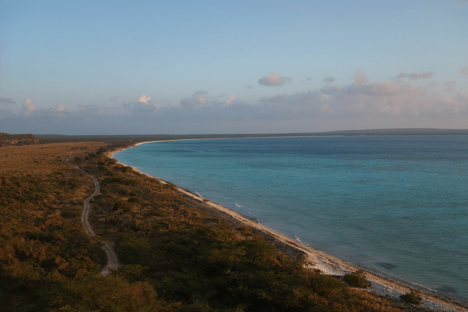 Bahía de las Águilas in Pedernales at dusk, with a curving Caribbean shoreline, dry coastal vegetation and a rough access track.