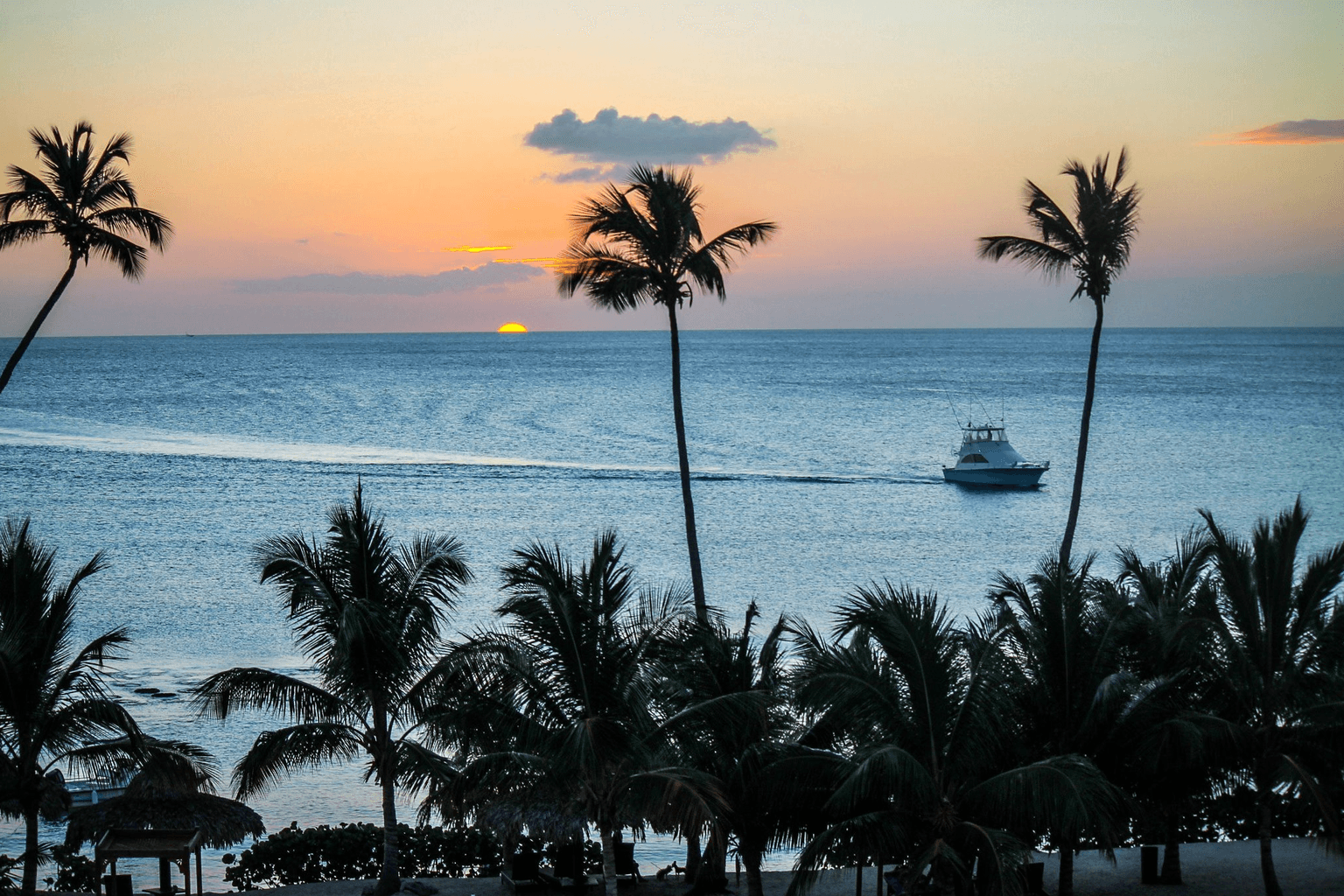 Sunset over Bayahíbe’s Caribbean coast with palm trees and a small boat crossing calm blue water.